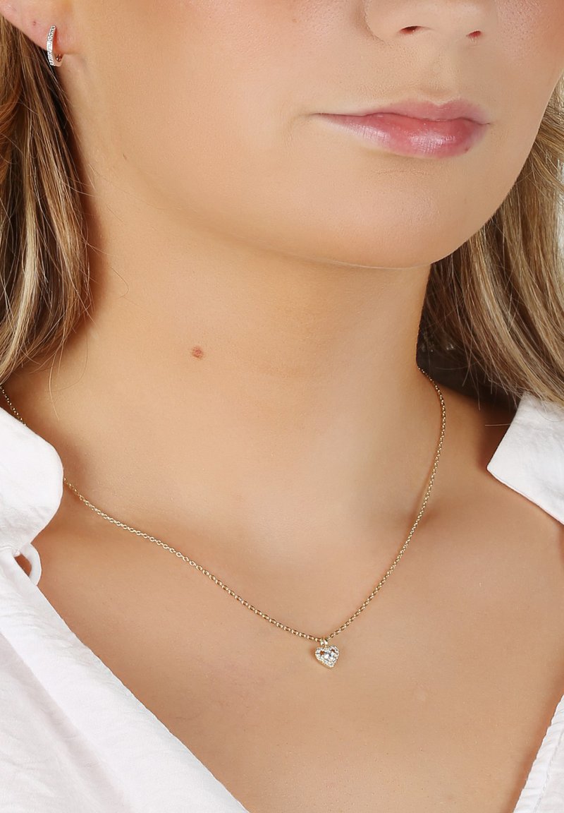 Guldhalsband med en liten hjärtformad hänge med funklande detalj. Bärs med en vit skjorta.