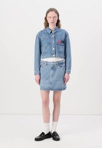 Kuttet denimjakke med to lommer og knappelukking, kombinert med et tettsittende denimskjørt. Begge i lyseblått, med synlige sømmer.