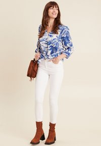 Blau-weiße Bluse mit Blumenmuster, weiße Skinny-Jeans, braune Wildleder-Stiefeletten und eine braune Fransen-Handtasche. Leger, mit lebhaften Mustern.