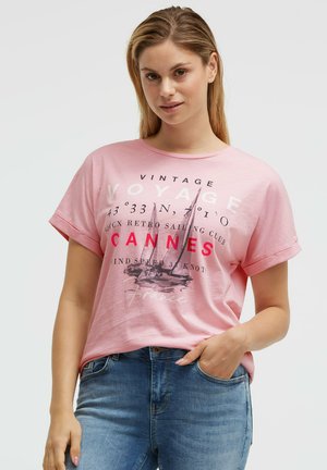 Frau mit langen blonden Haaren, die ein rosa Vintage-Segelclub-T-Shirt und blaue Jeans trägt, Hand in der Tasche, neutraler Hintergrund.