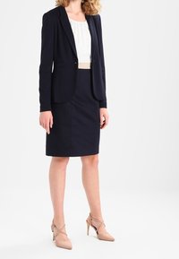 Kvinna klädd i en marinblå kavaj och matchande blyertskjol, vit blus och beige högklackade skor, stående mot en vit bakgrund.