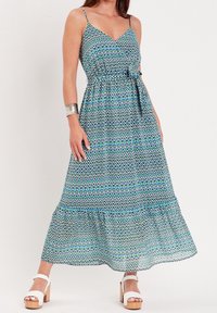 Robe longue avec un col en V, motif géométrique bleu et vert, fines bretelles spaghetti et taille cintrée par un lien. Portée avec des sandales blanches à talons blocs.