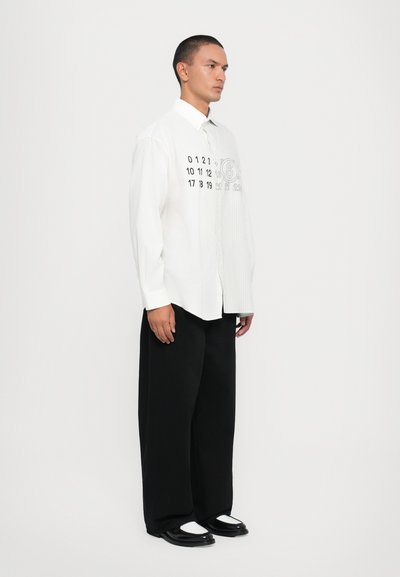 Weißes oversized Hemd mit schwarzen Zahlenprints auf einer Seite und vertikalen Streifen auf der anderen; kombiniert mit lockeren schwarzen Hosen und Schuhen.