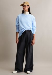 Lichtblauwe trui, zwarte wijde broek met zijstrepen, beige pet en witte schoenen; gelaagde coltrui zichtbaar onder de trui.