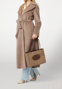 Beige lang jakke i imiteret pels med bælte, kombineret med en mønstret brun taske med logo. Denimflared bukser og ankelstøvler med slangeprint.