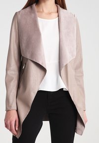 Chaqueta de imitación de cuero taupe claro con cuello de ante, apertura frontal y dos bolsillos laterales con cremallera. Llevada sobre una blusa blanca y pantalones negros.