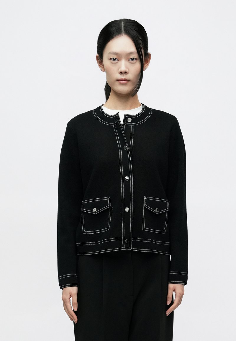 Fekete kardigán fehér varrással, kerek nyakkivágással, két elülső zsebbel és gombos lezárással. Az anyag puhanak tűnik enyhe text�úrával.