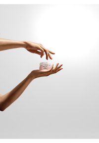 Szklany słoik z białą pokrywką, zawierający różowy krem, trzymany przez dwie jasnoskóre dłonie na neutralnym tle.