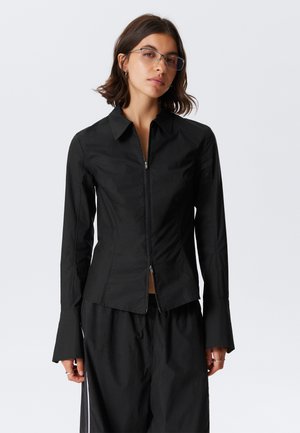 Young woman with shoulder-length hair wearing tinted glasses, a fitted black long-sleeve zip-up shirt, and loose black pants.