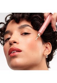 Różowozłota tubka z precyzyjnym aplikatorem trzymana blisko policzka; osoba ma gładką, promienną skórę, jasnoróżowe usta oraz krótkie, kręcone włosy.