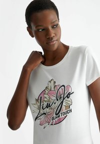 T-shirt bianca in cotone con un design grafico floreale e di scrittura colorato, con accenti rosa, neri e beige; scollo rotondo, maniche corte.