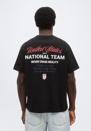 Άνδρας με κοντά σγουρά μαλλιά, ο οποίος φαίνεται από πίσω, φοράει μαύρο μπλουζάκι με το κείμενο «United States Men's National Team» και ένα λογότυπο των ΗΠΑ σε κόκκινο, άσπρο και μπλε.