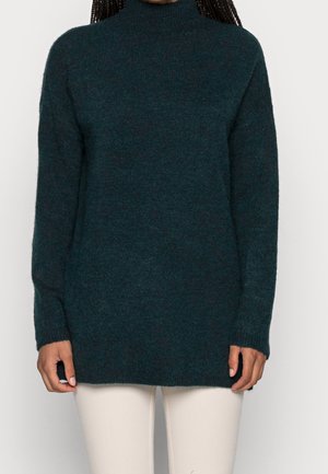 Donna che indossa un maglione verde scuro a maniche lunghe con collo alto abbinato a pantaloni beige chiaro, in piedi davanti a uno sfondo semplice.