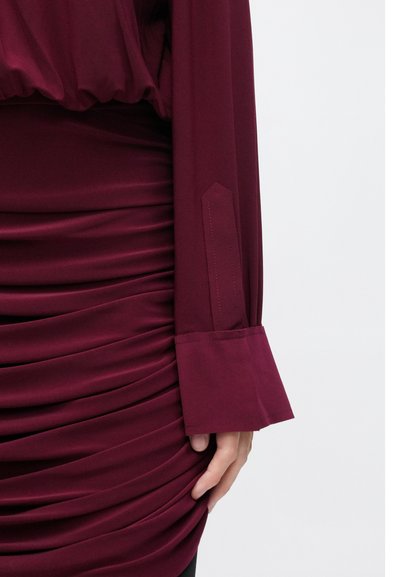 Bordo spalvos viršus su susmulkinta tekstūra ties juosmeniu, ilgomis rankovėmis su plačiomis rankogaliais ir aptempta suknele su vertikaliomis klostėmis.