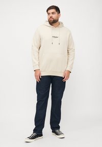 Beige Kapuzensweatshirt mit einer Fronttasche und schwarzem Logo, kombiniert mit dunkelblauen Jeans und schwarzen Turnschuhen. Lässiger Schnitt, weicher Stoff.