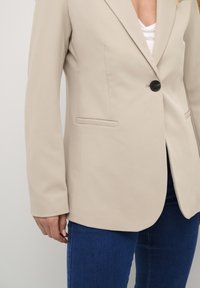 Béžový blazer s jedním černým knoflíkem, klopovými límci a dvěma předními kapsami, v kombinaci s modrými džíny. Hladká tkanina se strukturovaným tvarem.