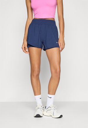 Marine sportshorts met een elastische tailleband en zijkanten, gecombineerd met een roze crop top en witte sneakers met donkere accenten.