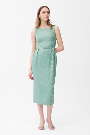 Vrouw draagt een mouwloze, muntgroene, knielange jurk met textuur en een aansluitende taille, staand tegen een effen witte achtergrond, kijkt opzij.