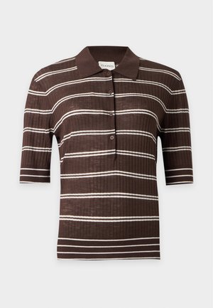 Femme portant un polo à manches courtes rayé brun et blanc, un pantalon gris clair et des ballerines noires, debout devant un fond uni.