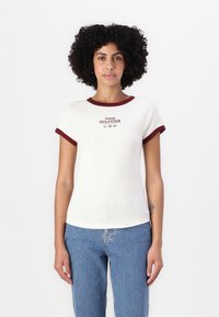 Bela majica s kratkimi rokavi z grimiznim ovratnikom in rokavi, ki nosi logotip "Tommy HILFIGER" v sredini v temno rdeči barvi. Mehka bombažna tkanina.