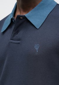 Pánske polo tričko v námorníckej modrej farbe s ryhovanou textúrou a tmavomodrým golierom. Na hrudi je malý vyšívaný dizajn v tvare srdca.