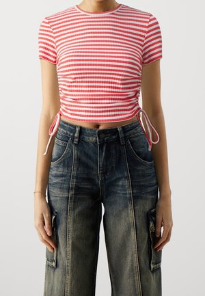 Femme portant un top court rayé rouge et blanc avec des liens sur les côtés et un jean cargo noir délavé taille haute, debout devant un fond uni.