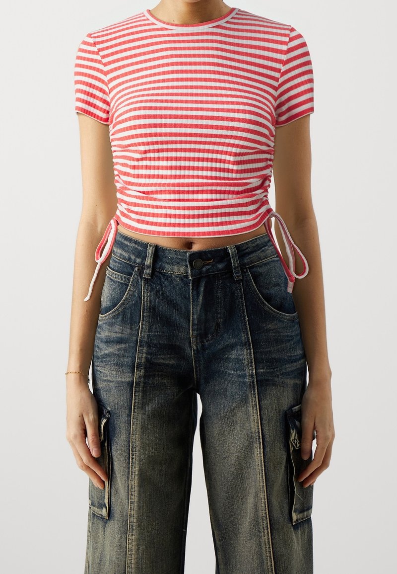 Femme portant un top court rayé rouge et blanc avec des liens sur les côtés et un jean cargo noir délavé taille haute, debout devant un fond uni.