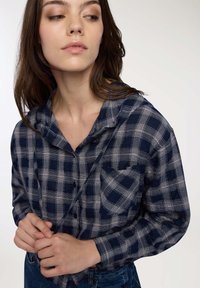 Giovane donna con lunghi capelli castani, che indossa una camicia a quadri blu navy e grigia, mentre regola il cordino della camicia guardando di lato.
