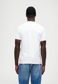 Uma t-shirt branca de manga curta feita de algodão, com um decote redondo e design sem costuras, utilizada com jeans azuis. O tecido parece suave.