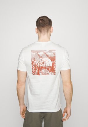 Person iført hvid T-shirt med orange bjerg- og skovgrafik samt "The North Face"-logo på ryggen, stående mod en ensartet baggrund.