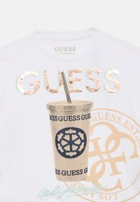 Λευκό βαμβακερό μπλουζάκι με κεντητό σχέδιο κυπέλλου, κείμενο σε χρυσό και μπλε χρώμα, και μεταλλικές λεπτομέρειες. Το σχέδιο περιλαμβάνει κυκλικό μοτίβο λογότυπου.