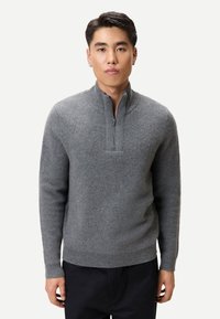 Junger Mann, der einen grauen Pullover mit halbem Reißverschluss und schwarze Hose trägt, steht vor einem schlichten hellen Hintergrund und blickt geradeaus.