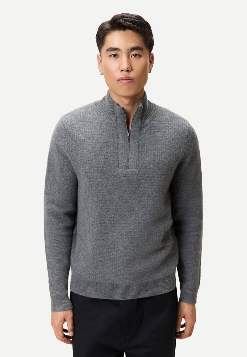 Junger Mann, der einen grauen Pullover mit halbem Reißverschluss und schwarze Hose trägt, steht vor einem schlichten hellen Hintergrund und blickt geradeaus.