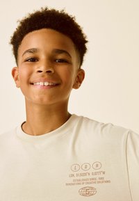Beige t-shirt med ribbad halsringning, med bruna textgrafik på framsidan, inklusive koordinater och varumärkesdetaljer.