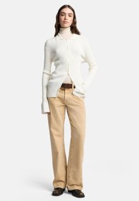 Biały prążkowany kardigan na guziki, noszony na jasnym golfie, zestawiony z jasnobeżowymi rozkloszowanymi spodniami i brązowymi butami. Gładkie tekstury.