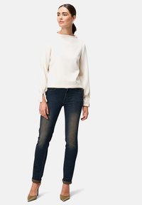 Lichtcrème top met hoge hals en lange mouwen, gecombineerd met donkerblauwe skinny jeans en beige hoge hakken.