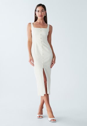 Fitted light cream dress with square neckline, front button detailing, and thigh-high slit. Made of smooth fabric. Worn with white heels.