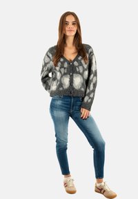 Grijze en witte gebreide cardigan met een V-hals, knoopsluiting en lange mouwen, gecombineerd met blauwe jeans en beige sneakers.