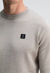 Beige gebreide trui met een kleine zwarte patch waarop een gestileerde witte "J" staat op de bovenste borst, gedragen door een persoon met een lichte huid.