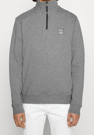 Homme portant un pull gris à fermeture éclair au quart avec le logo "BOSS" sur la poitrine et un pantalon blanc, debout devant un fond uni.