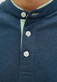 Poloshirt aus dunkelblauem, strukturiertem Stoff mit einem grünen Kragen. Verfügt über eine kurze Knopfleiste mit drei weißen Knöpfen und einer Brusttasche.