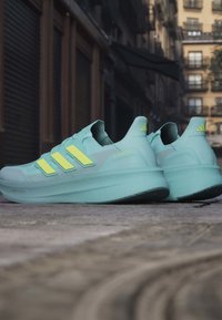 Helles blaues Sportschuhpaar mit drei leuchtend gelben Streifen, strukturiertem Obermaterial und einer dicken, gepolsterten Sohle mit schwarzem Gummiprofil.