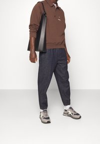 Polo a maniche lunghe marrone, pantaloni in denim scuri, sneakers grigie e navy, e una borsa tote nera; tessuto testurizzato e vestibilità rilassata.