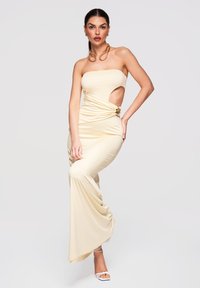 Femme portant une robe beige bustier avec une découpe latérale et une boucle dorée, posant avec une main sur la hanche et des sandales blanches à talons.