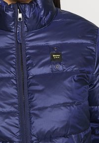 Doudoune marine avec un col montant, fermeture éclair YKK, coutures horizontales et une étiquette en tissu noir avec le logo "Blauer U.S.A.".