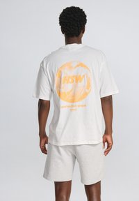 Hvidt bomulds T-shirt med orange grafik med teksten "HSW" og "Serve the game", parret med lys grå shorts. Enkel, afslappet pasform.