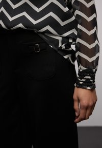 Černobílá halenka s chevron vzorem, vyrobená z průsvitného materiálu, s černými volánovými manžetami, v kombinaci s černými kalhotami s boční kapsou a detaily opasku.