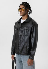 Zwarte nepleren jas met knoopsluiting aan de voorkant, twee borstzakken en zichtbare stikdetails. Model draagt lichtblauwe spijkerbroek en gele zonnebril.