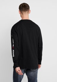 Svart långärmad T-shirt med rund hals, med vit text "TOMMY JEANS" på vänster ärm. Bomullsstruktur, avslappnad passform.