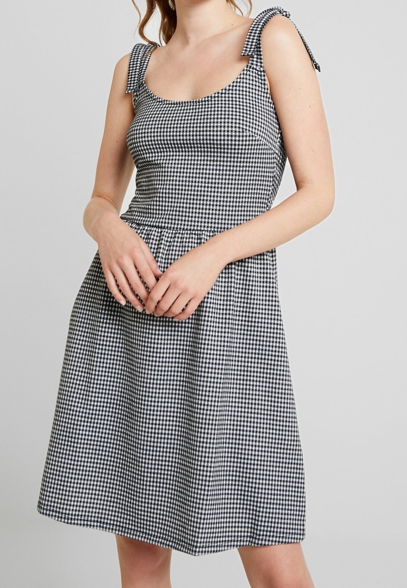 Femme portant une robe sans manches à carreaux vichy noir et blanc avec des bretelles nouées aux épaules, debout devant un fond clair uni.